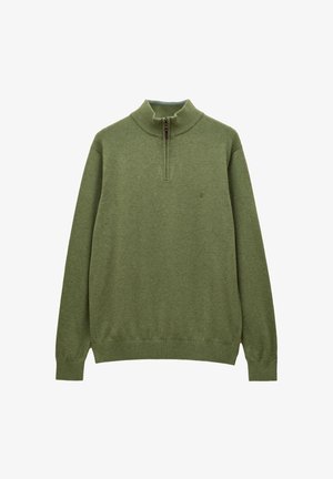 Mann trägt grünen Viertel-Reißverschluss-Pullover über kariertem Hemd und beigen Hosen, steht mit Händen in den Taschen vor einfarbigem beigem Hintergrund.