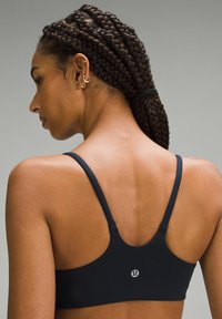 Čierna športová podprsenka s nastaviteľnými tenkými ramienkami, hladkou textúrou a logom na chrbte. Viditeľná pleť s vrkôčkami.
