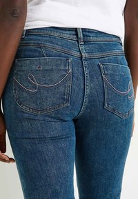 Pantalones vaqueros de mezclilla en azul oscuro con un ligero desgaste, que cuentan con bolsillos traseros cosidos y un diseño de cintura alta. Textura y ajuste consistentes.