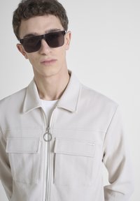Chaqueta de cremallera en un tono beige claro con dos bolsillos en el pecho y detalles en hardware plateado. Luce sobre una camisa blanca, combinada con gafas de sol oscuras.