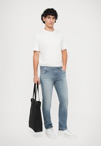 Ανοιχτό μπλε τζιν παντελόνι με εφαρμογή slim fit, συνδυασμένο με λευκή μπλούζα και μαύρη τσάντα tote. Τα λευκά αθλητικά παπούτσια ολοκληρώνουν την εμφάνιση.