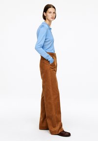 Frau steht seitlich in einem hellblauen Langarmshirt, weiten braunen Cordhosen mit Taschen und dunkelbraunen Schuhen vor einem weißen Hintergrund.