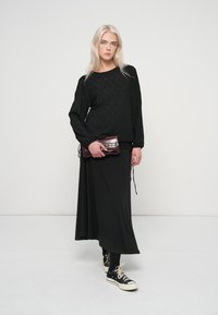 Μαύρο μακρυμάνικο τοπ με υφή, συνδυασμένο με μακριά μαύρη φούστα, κρατώντας ένα σκούρο κόκκινο τσαντάκι. Μαύρα αθλητικά παπούτσια με λευκές σόλες.