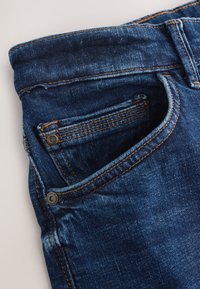 Gros plan d’un jean en denim bleu montrant la poche avant, la petite poche à monnaie, la passants de ceinture et les détails de couture orange.