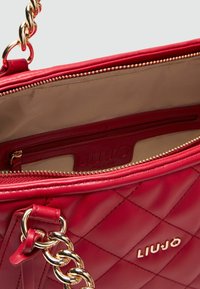 Rote gesteppte Handtasche mit einer goldenen Ketten-Tragriemen und Reißverschluss. Das Innenfutter hat einen glatten beigen Stoff mit einer Reißverschlusstasche und einem Markenetikett.