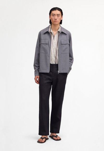 Mladý muž ve šedé bundě na zip, pruhované košili na knoflíky, černých kalhotách a černých žabkách na jednoduchém bílém pozadí.