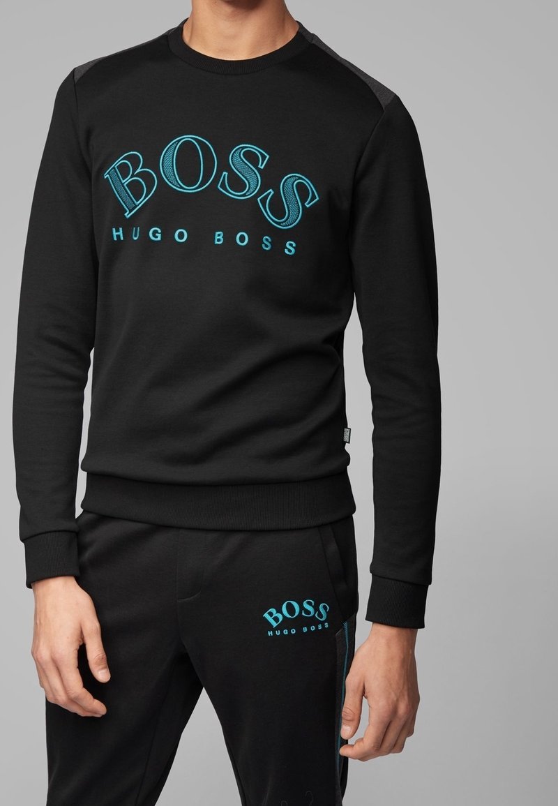 Sudadera negra de material suave con logo "BOSS" en turquesa y texto "HUGO BOSS"; puños y bajo acanalados; combinada con pantalones a juego.