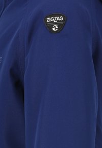 Marineblauwe stoffen jas met een zwarte driehoekige patch met het label "ZIGZAG" op de linkerschouder, met zichtbare stikseldetails.