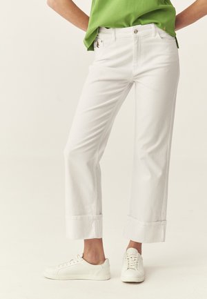 Personne portant un pantalon blanc retroussé, des baskets blanches et une chemise verte, debout contre un fond clair uni.