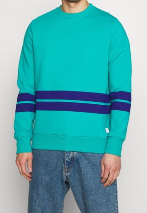 Persoon draagt een teal sweatshirt met donkerblauwe horizontale strepen op de mouwen en romp, gecombineerd met blauwe spijkerbroek.