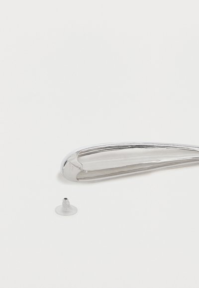 Μακριά, καμπυλωτή ασημένια σκουλαρίκι με μικρό διαχωρισμένο διαφανές πλαστικό πίσω μέρος πάνω σε λευκή επιφάνεια.