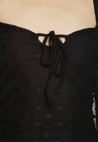 Mustat yläosa, jossa on pilkullinen rakenne, neliömäinen pääntie ja solmukohta keskellä. Valmistettu läpikuultavasta materiaalista.