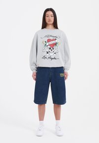 Harmaa huppari, jossa on värikäs pääkallo grafiikka tekstillä, yhdistettynä polvenpituisiin sinisiin farkkushortseihin, joissa on keltainen tekstiyksityiskohta, sekä valkoisiin lenkkareihin.