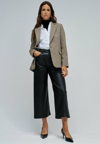 Una mujer de pie vestida con un blazer beige, una camisa blanca con cuello de tortuga negro, pantalones de cuero negros de pierna ancha y tacones de punta negros, contra un fondo gris.