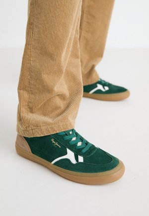 Zöld velúr sportcipő fehér díszítéssel és zöld fűzőkkel. Gumitalp, kerek orr és bézs részletek a sarokon. Csíkos nadrággal viselve.