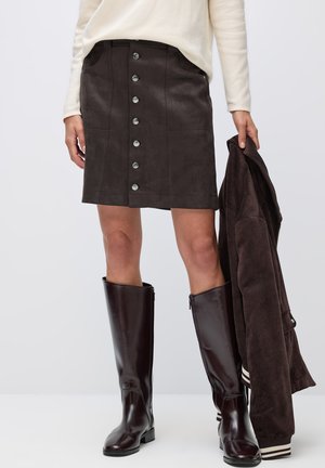 Jupe en suède marron avec fermeture à boutons sur le devant, poches latérales et texture lisse. Associée à des bottes en cuir marron montant jusqu'aux genoux.