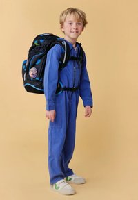 Combinaison bleue avec fermeture éclair, sac à dos noir avec des accents bleus et un badge circulaire, et chaussures blanches avec des détails verts.