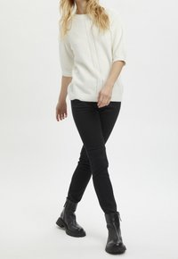 Maglione bianco lavorato a maglia con un design testurizzato, abbinato a pantaloni neri slim-fit e stivaletti neri, che mostrano un outfit casual.