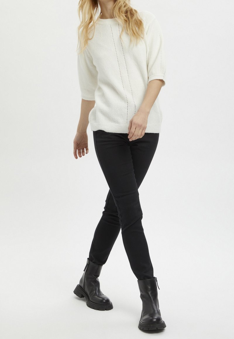 Maglione bianco lavorato a maglia con un design testurizzato, abbinato a pantaloni neri slim-fit e stivaletti neri, che mostrano un outfit casual.