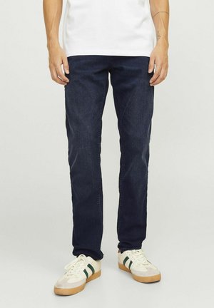Homme portant un jean slim bleu foncé, des baskets blanches avec des bandes vertes et une chemise blanche, debout devant un fond uni.