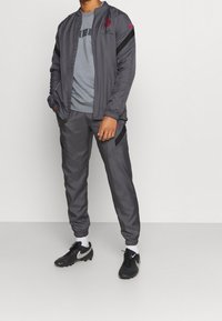 Completo sportivo grigio composto da una giacca con zip e pantaloni affusolati con polsini in elastico. Accenti rossi e dettagli del logo sul tessuto.
