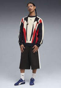 Uomo con capelli intrecciati che indossa un maglione Puma rosso, nero e bianco, pantaloncini larghi neri, calzini bianchi e sneakers Puma blu, in piedi davanti a uno sfondo grigio.