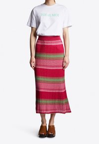 Vit t-shirt med "TOI et MOI" i grönt, tillsammans med en veckad kjol i röd och rosa randig design med gröna accenter och bruna trätofflor.