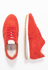 Punaiset mokkanahkakengät, joissa on pyöreä kärki, nauhakiinnitys, vaaleanruskea kumipohja ja valkoinen välikerros. Sivussa on ompeluskoristeita.