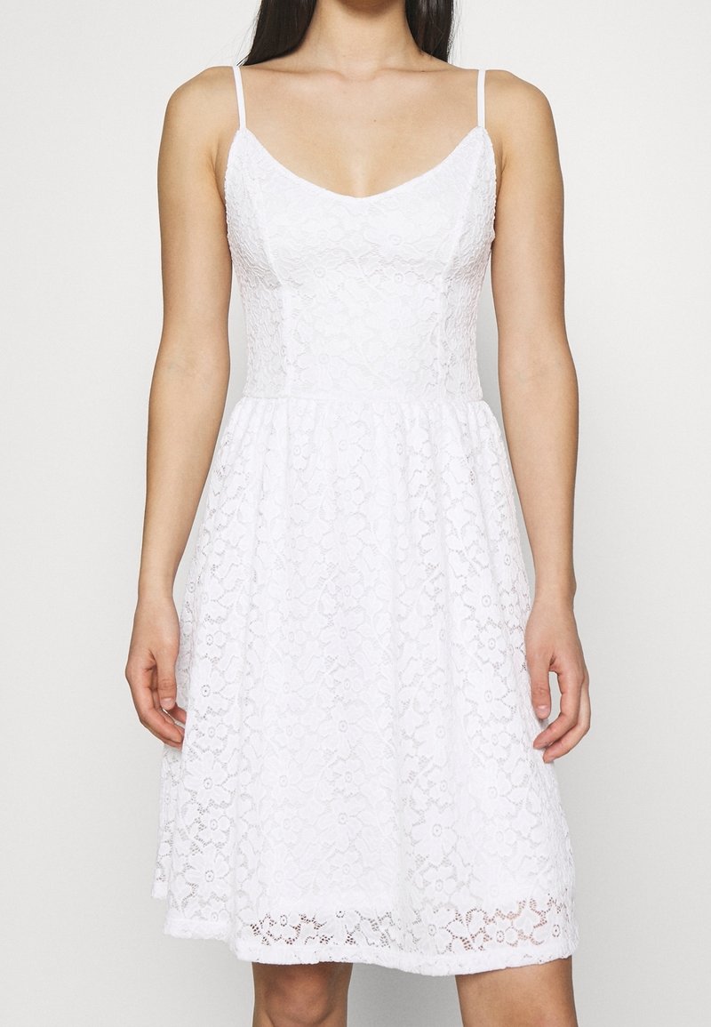 White lace dress with adjustable spaghetti straps, a fitted bodice, and a flared skirt. Floral pattern throughout and scalloped hem detailing.