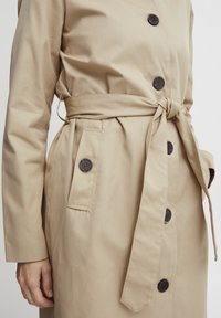 Beiger Trenchcoat mit Gürtel in der Taille, langen Ärmeln, zwei seitlichen Taschen und dunklen Knöpfen. Glatte Textur und maßgeschneiderte Design.