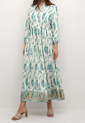 Vrouw die een lange witte jurk met teal bloemenpatronen draagt en beige sandalen met blokhak en open tenen, staand tegen een effen achtergrond.