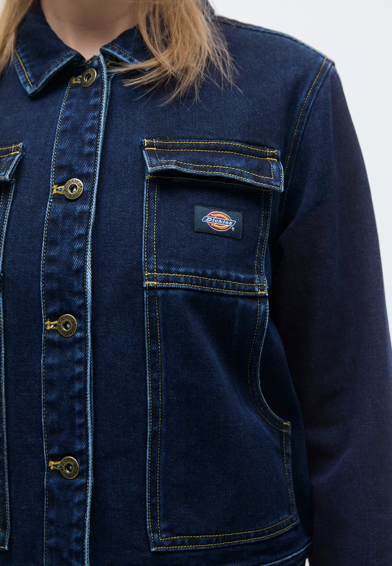 Denimjack in donkerblauw met gouden stiksels. Heeft een knoopsluiting, twee borstzakken en een merkpatch aan de linkerkant.