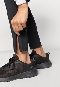 Juodi sportiniai džinsai su šonine užtrauktuko detale, derinami su juodais orui pralaidžiais sportbačiais ir tekstūruota nitinimo danga.