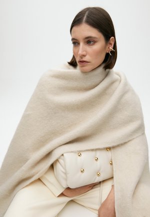 Beige Wollmischung-Schal hängt über einer cremefarbenen, weichen Clutch mit ausgeprägten goldenen Nieten. Das Modell posiert mit sanft zusammengelegten Händen um die Tasche.