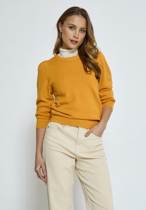 Mosterdgele gebreide trui met een ronde hals, geribbelde manchetten en zoom; gecombineerd met lichtbeige wijde broek.