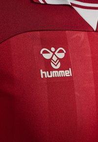 Röd träningströja med en framträdande vit "hummel"-logotyp och en design av en bi. Tyget har en slät textur och en subtil glans.