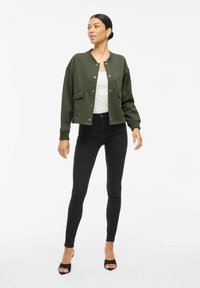 Zaļa kroptā bomber jaka ar apaļo apkakli, spiedpogu aizdari un priekšējām kabatām, kombinēta ar melniem pieguļošajiem džinsiem un atvērtām papēža kurpēm.