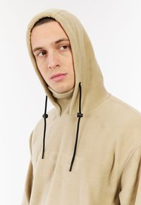 Beige fleece hoodie met een verstelbare capuchon, zwarte accenten en een reliëflogo op de borst. Zachte textuur en een relaxte pasvorm.
