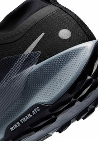Zapatilla Nike Trail ATC en negro y gris, con un upper texturizado, acentos reflectantes y una suela de goma con un patrón y surcos profundos.