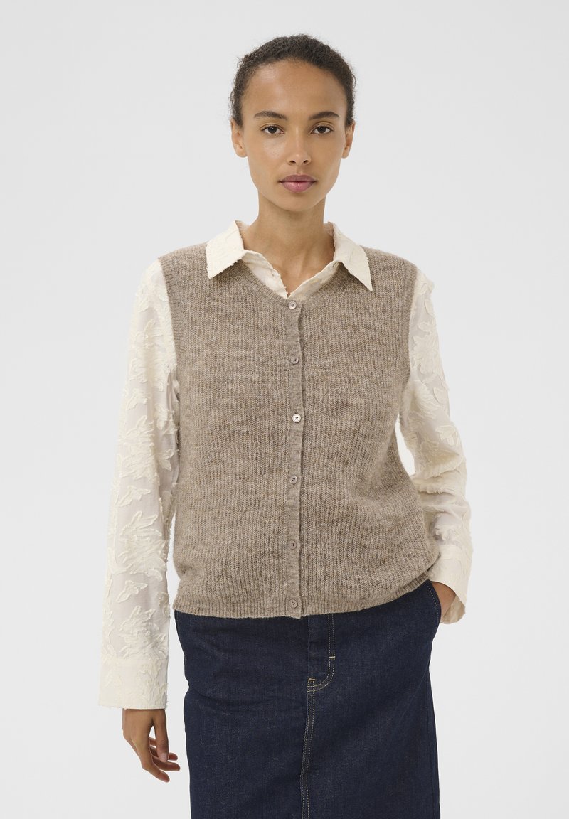 Beige strikvest med v-hals, knaplukning foran og ribbet tekstur, lagdelt over en hvid, broderet skjorte med lange ærmer. Mørk denimnederdel.