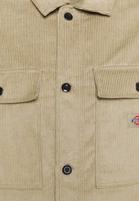 Giacca in corduroy beige con chiusura a bottoni, due tasche frontali e bottoni neri. Include una piccola etichetta logo sul lato sinistro.