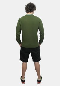 Maglietta verde oliva a maniche lunghe con collo rotondo, abbinata a pantaloni corti neri e scarpe sportive con dettagli arancioni. Vista posteriore mostrata.