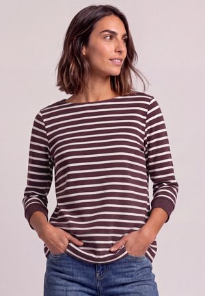 Femme aux cheveux mi-longs portant un t-shirt à manches longues rayé marron et blanc et un jean bleu, les mains dans les poches, regardant vers la droite.
