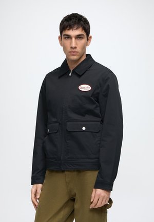 Homme portant une veste noire zippée avec col et deux poches avant à rabat, associée à un pantalon vert olive, debout devant un fond uni.