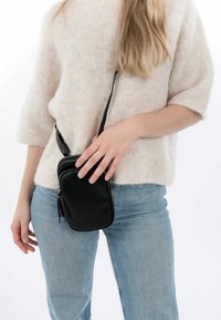 Schwarze Leder-Crossbody-Tasche mit zwei Reißverschlussfächern, glatter Textur und verstellbarem Riemen, in einer Hand gegen einen hellen Hintergrund gehalten.