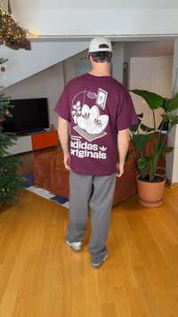 Homme vu de dos portant un t-shirt bordeaux Adidas Originals, un pantalon gris, une casquette blanche, debout dans un salon avec un sapin de Noël décoré.