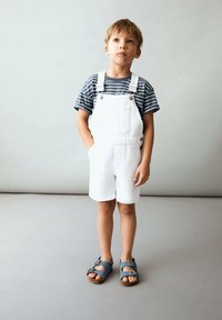 Weiße Denim-Latzhosen mit kurzen Beinen, gestreiftes T-Shirt in Marineblau und Weiß sowie graue Sandalen mit Schnallen. Einfaches Design, keine Muster oder Grafiken.