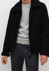 Sort faux suede jakke med en tekstureret sort shearling krave og manchetter, frontlynlås og sidelommer. Båret over en grå sweater.