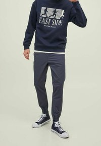 Uomo con indosso una felpa blu navy "East Side New York Runners", pantaloni jogger scuri e sneakers alte blu navy con lacci bianchi.