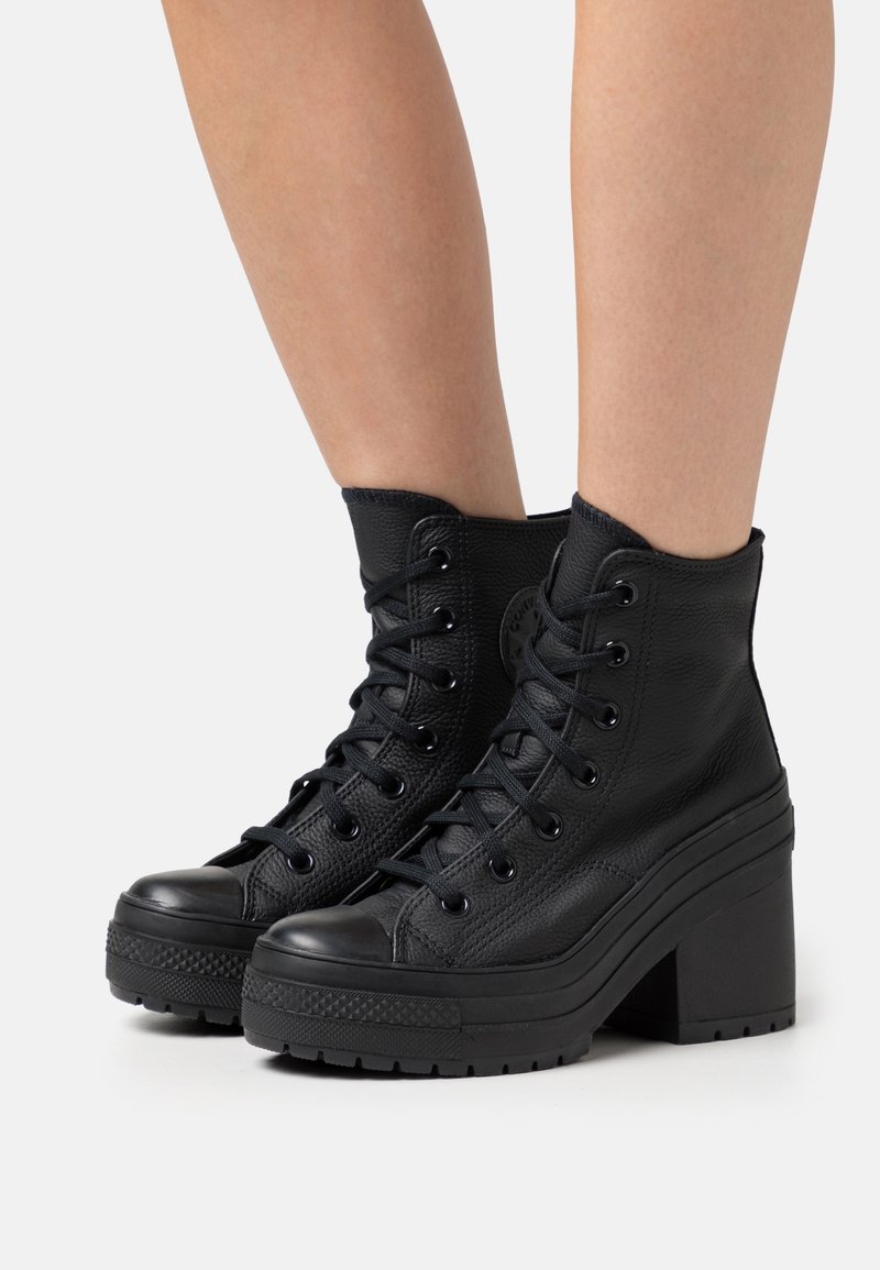 Bottines noires en cuir à talons hauts avec lacets, portées sur jambes nues, avec semelles épaisses à plateforme et bouts texturés.